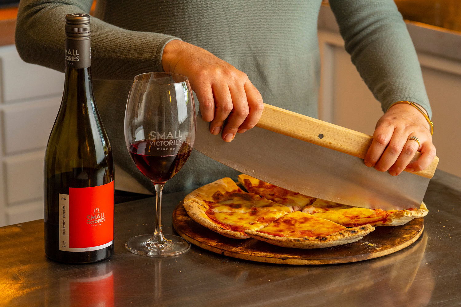Person slicing pizza on a pizza stone with a bottle and glass of Small Victories Sangiovese red wine beside it.