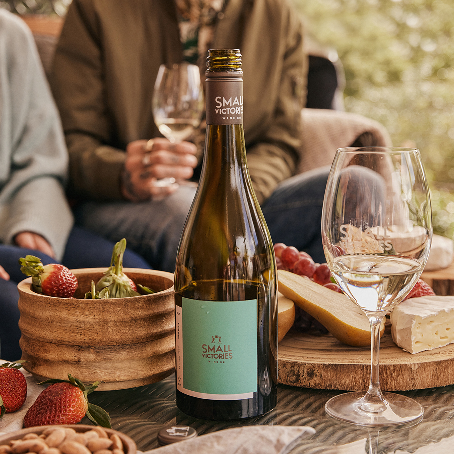 Small Victories Pinot Gris bottle on a table with strawberries, almonds, cheese, and grapes; two people in the background holding wine glasses.