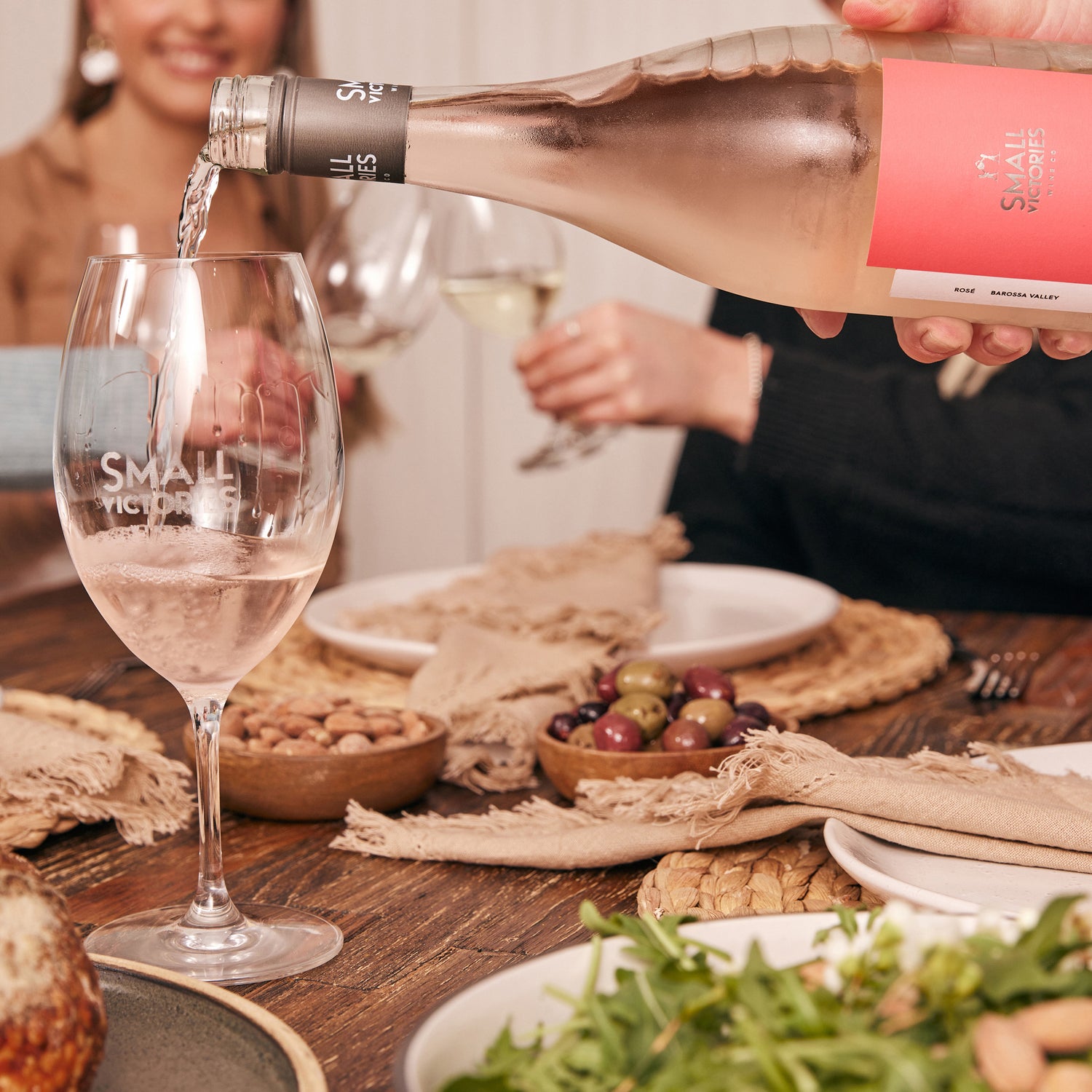 Close-up of a dining table with mixed olives and almonds, as wine is poured from a Small Victories Rose bottle into a branded glass; two people in the background holding wine glasses.