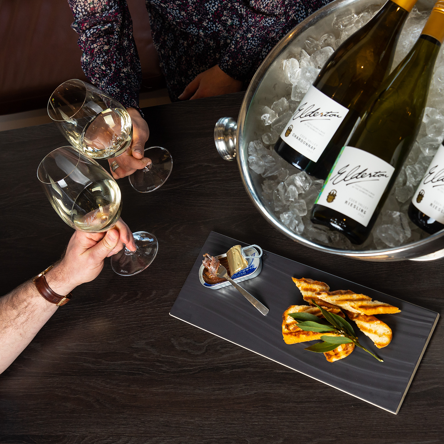 two hands cheersing 2 glasses of white wine, alongside wine in an icebucket and sardines and toast on a platter