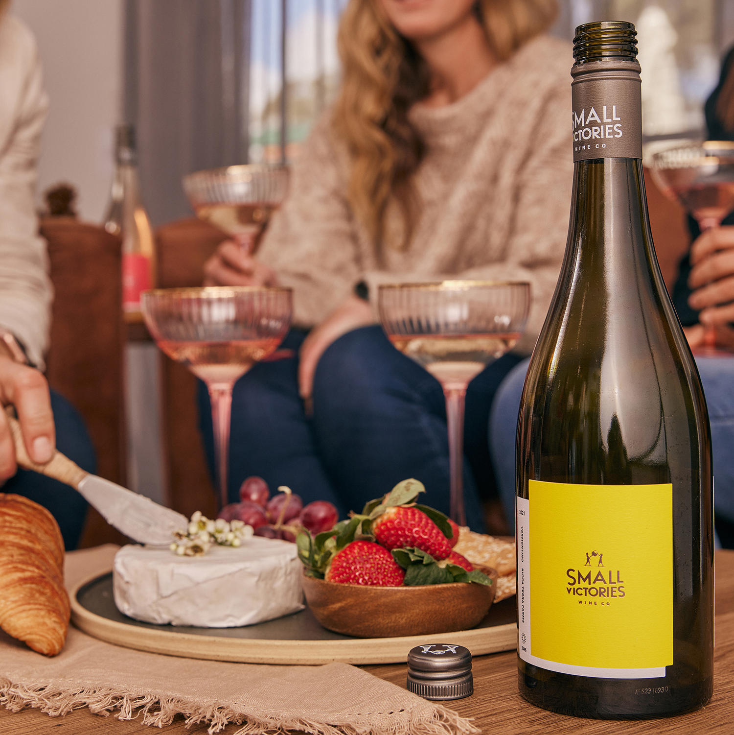 hand cutting a wheel of Camembert on a cheese plate with fruit, alongside a bottle of Small Victories Vermentino