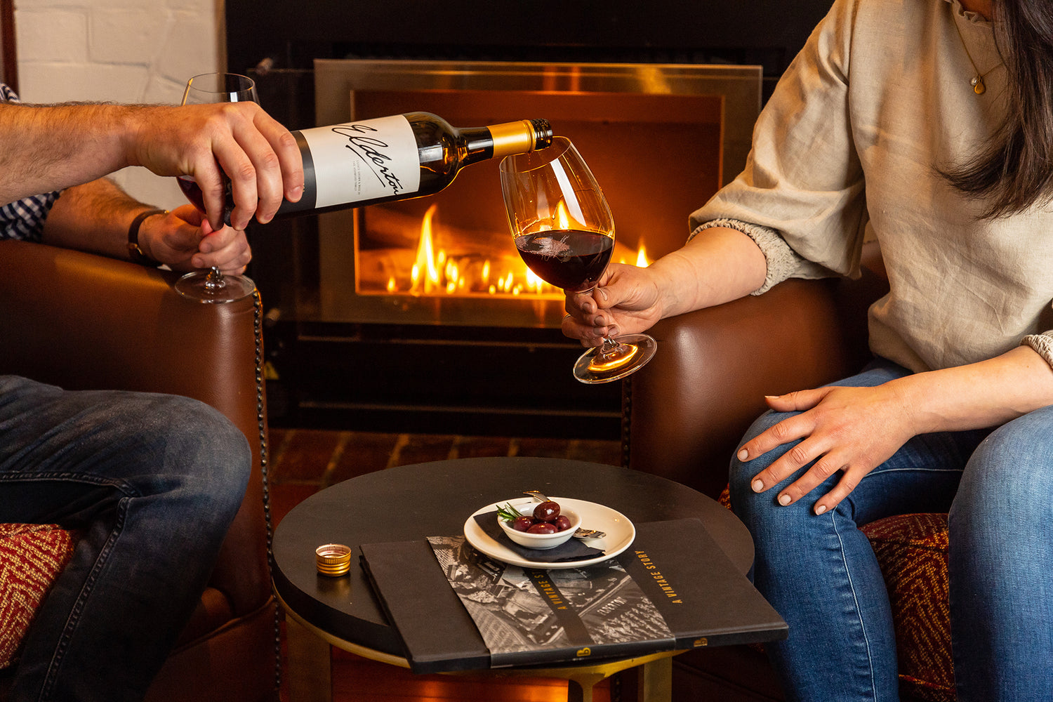 Two people sitting in armchairs with a glass of wine and a olives in front of a fireplace.